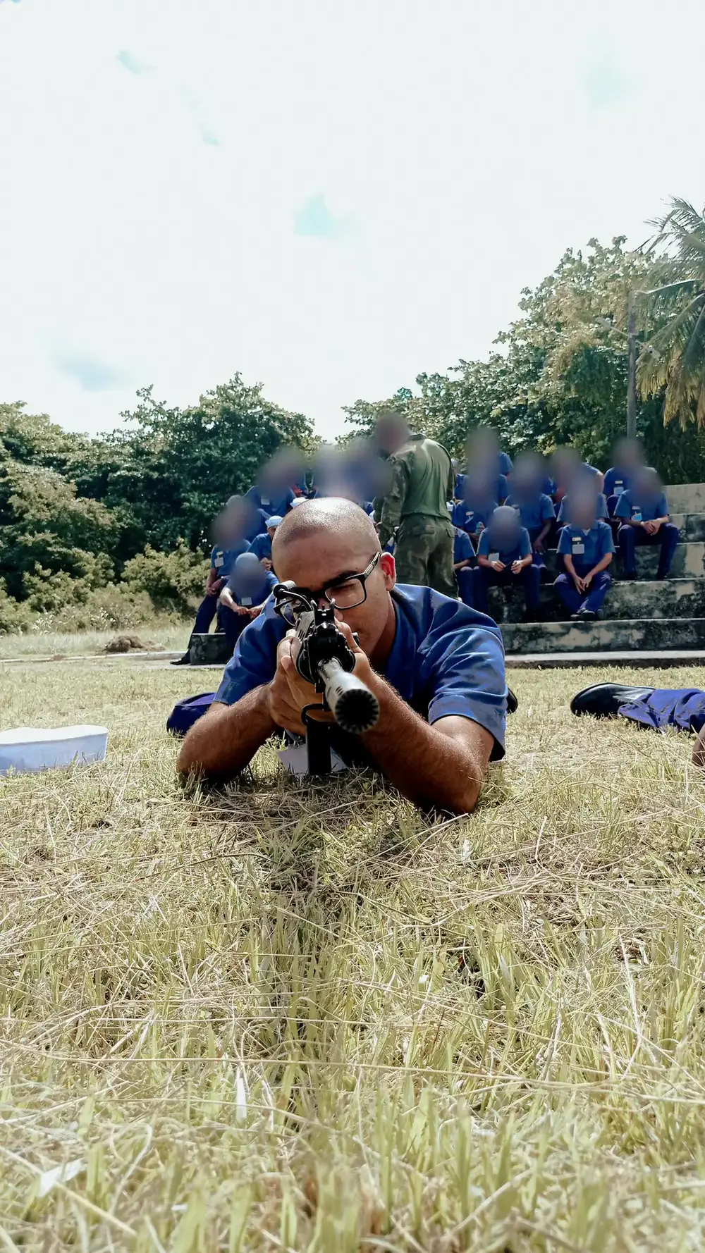 Daniel P Francisco deitado no gramado em posição de tiro durante um treinamento militar, com os rostos das pessoas ao fundo desfocados